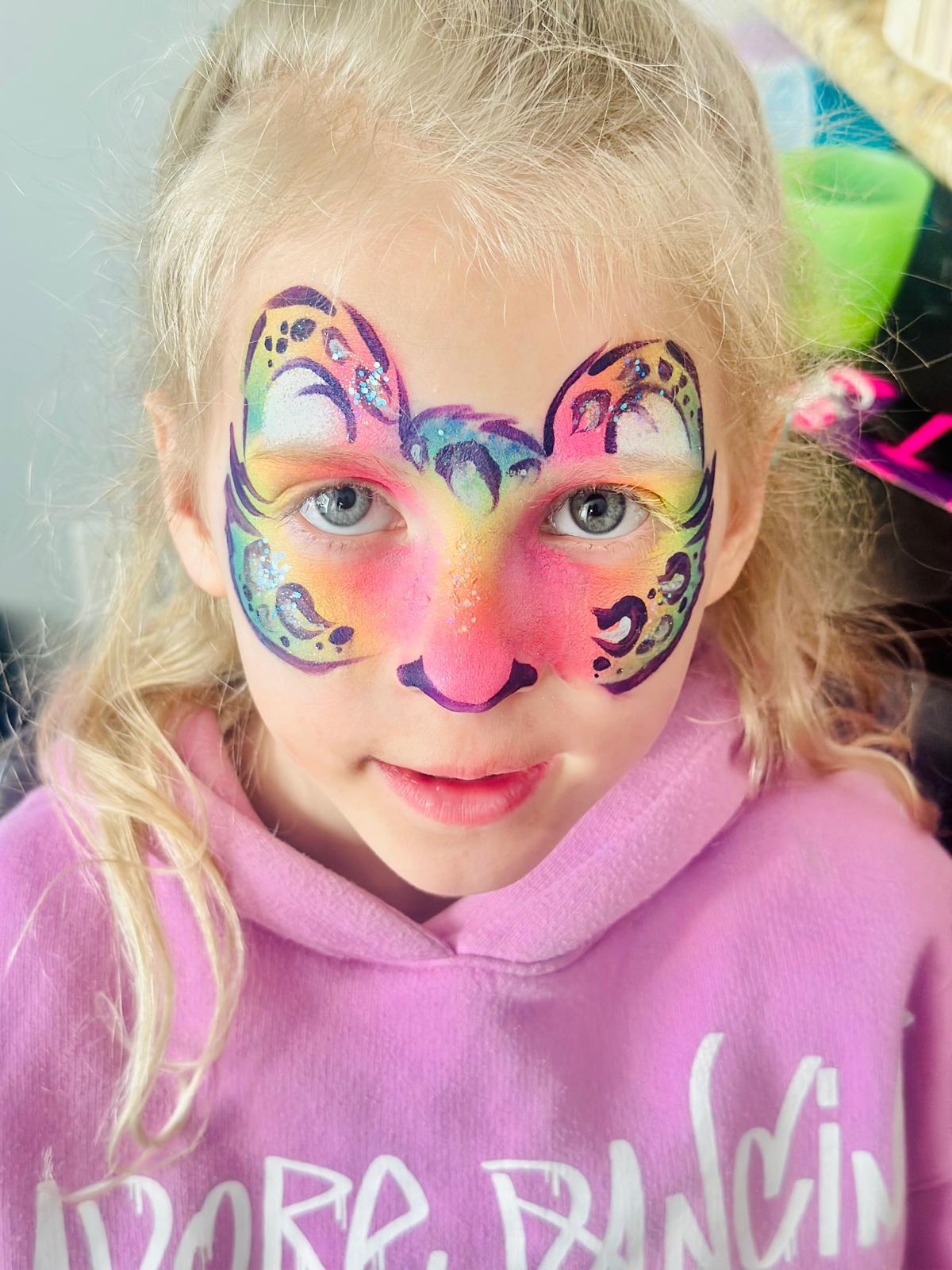 A young girl with a cat-themed face painting featuring pink, yellow, and blue hues