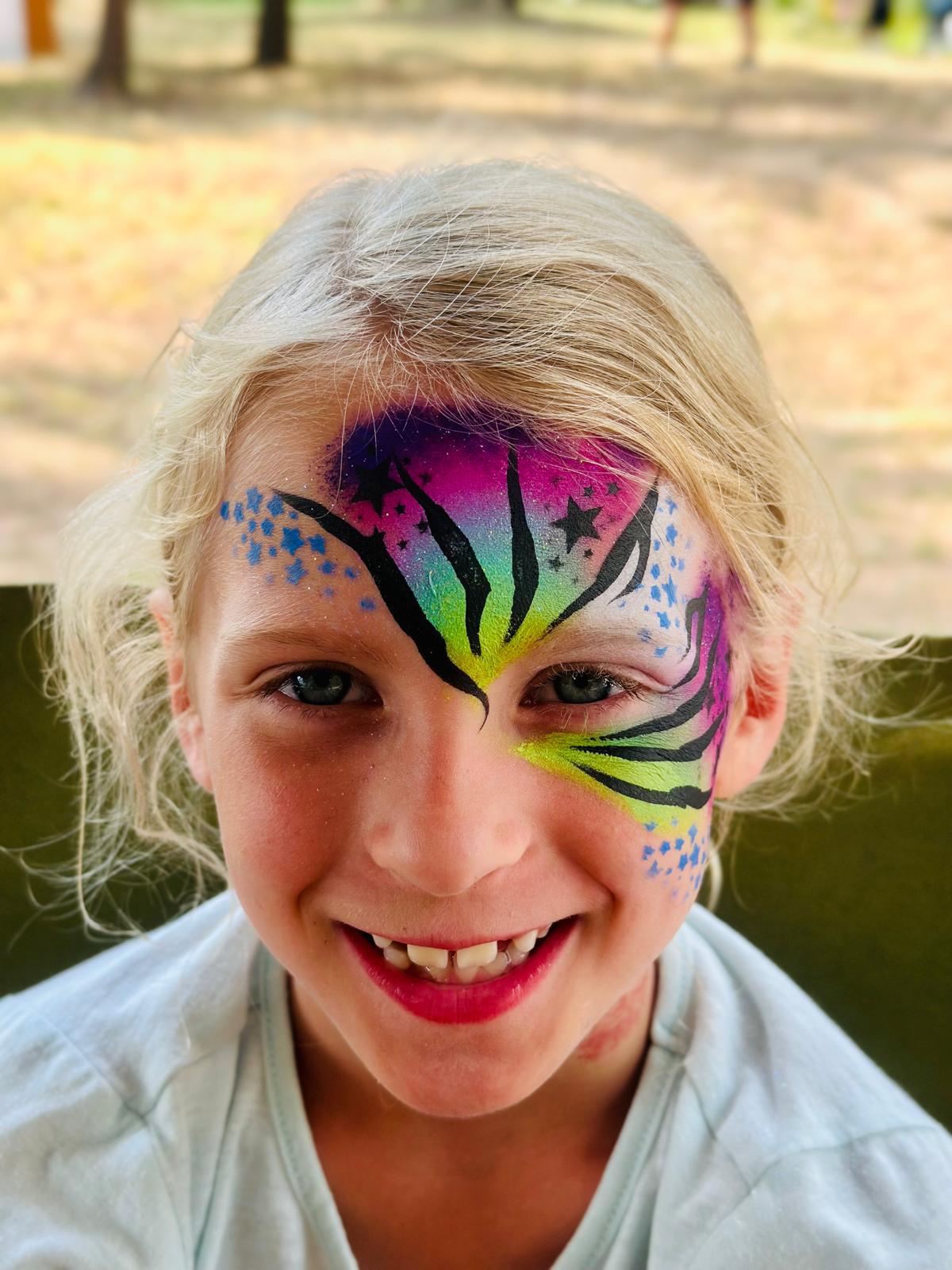 A smiling girl with a vibrant butterfly face painting in purple, green, and black