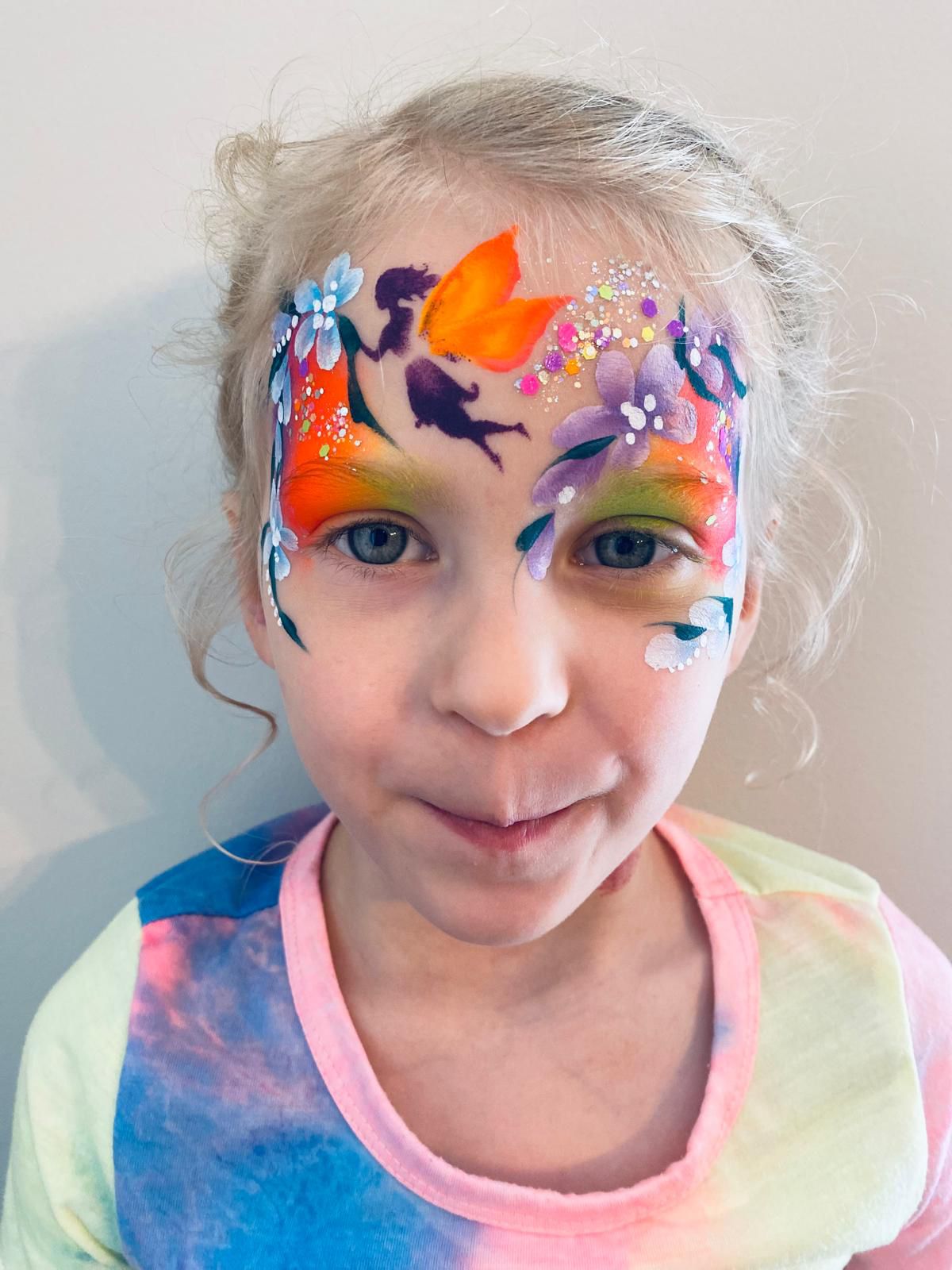 A young girl with an intricate face painting of a sunset scene featuring birds, flowers, and vibrant orange and purple shades